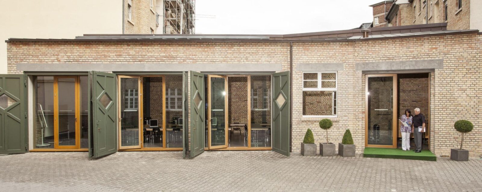 Brick facade of a building with open glass doors and two people under a canopy on the right side